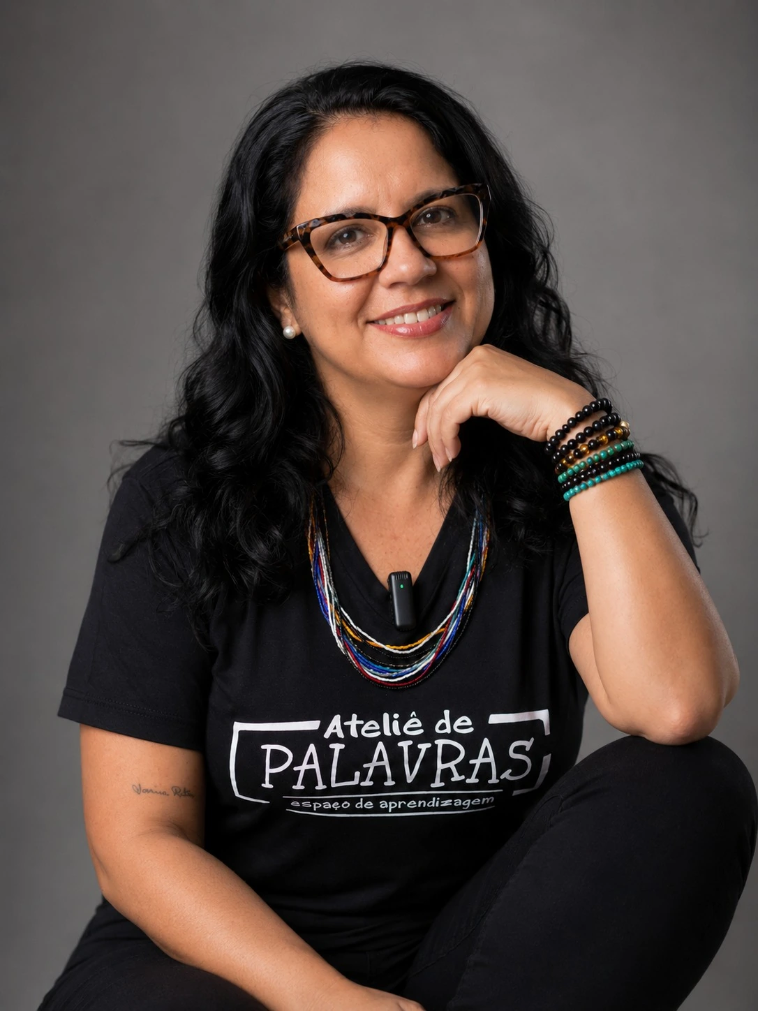 Retrato da Professora Bruna Valério, sorrindo, com óculos e camiseta preta com a estampa Ateliê de Palavras — espaço de aprendizagem