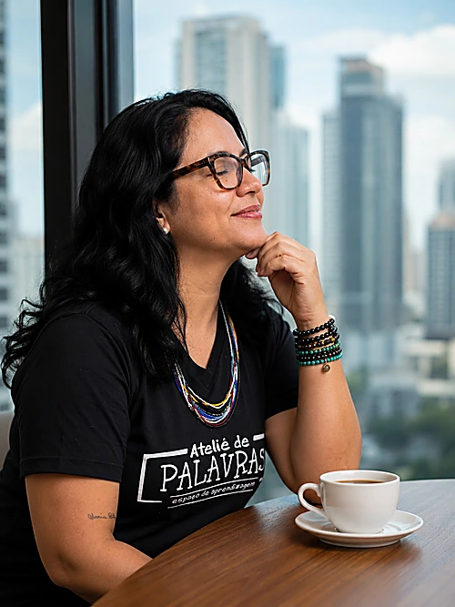 Professora Bruna Valério pensativa, olhando pela janela com uma xícara de café à frente, vestindo camiseta preta com a estampa Ateliê de Palavras